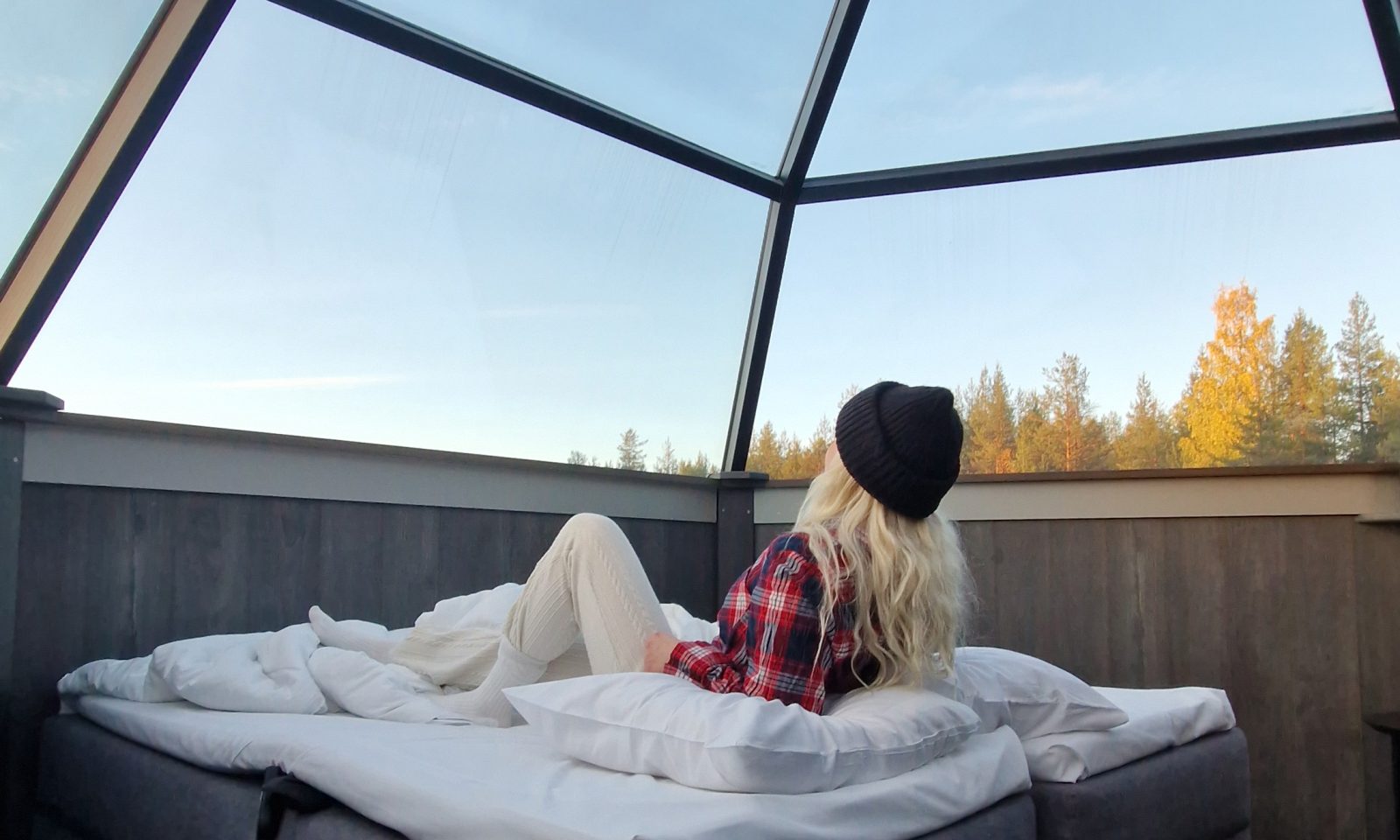 A person lying on a bed in a glass igloo and watching the autumn landscape